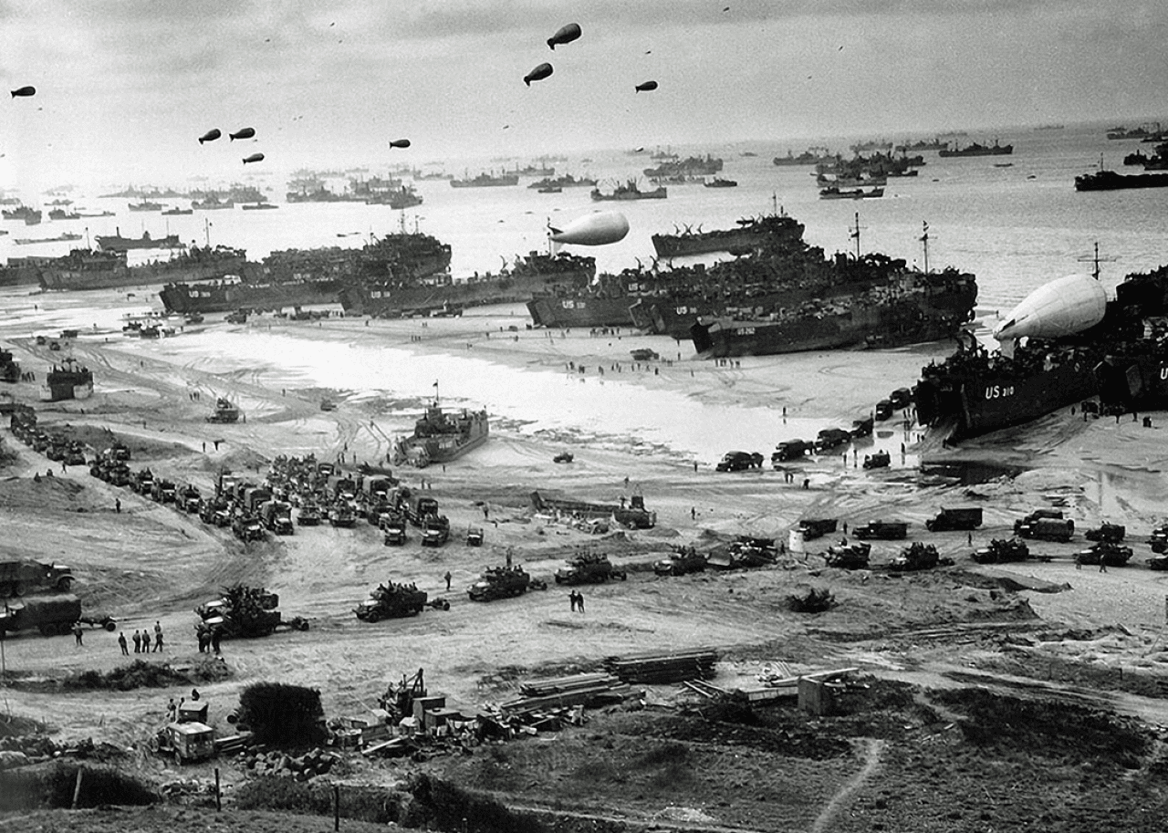 A large number of ships in front of Omaha Beach delivering troops, equipment and vehicles lining up to leave the beach, barrage balloons are up in the sky.